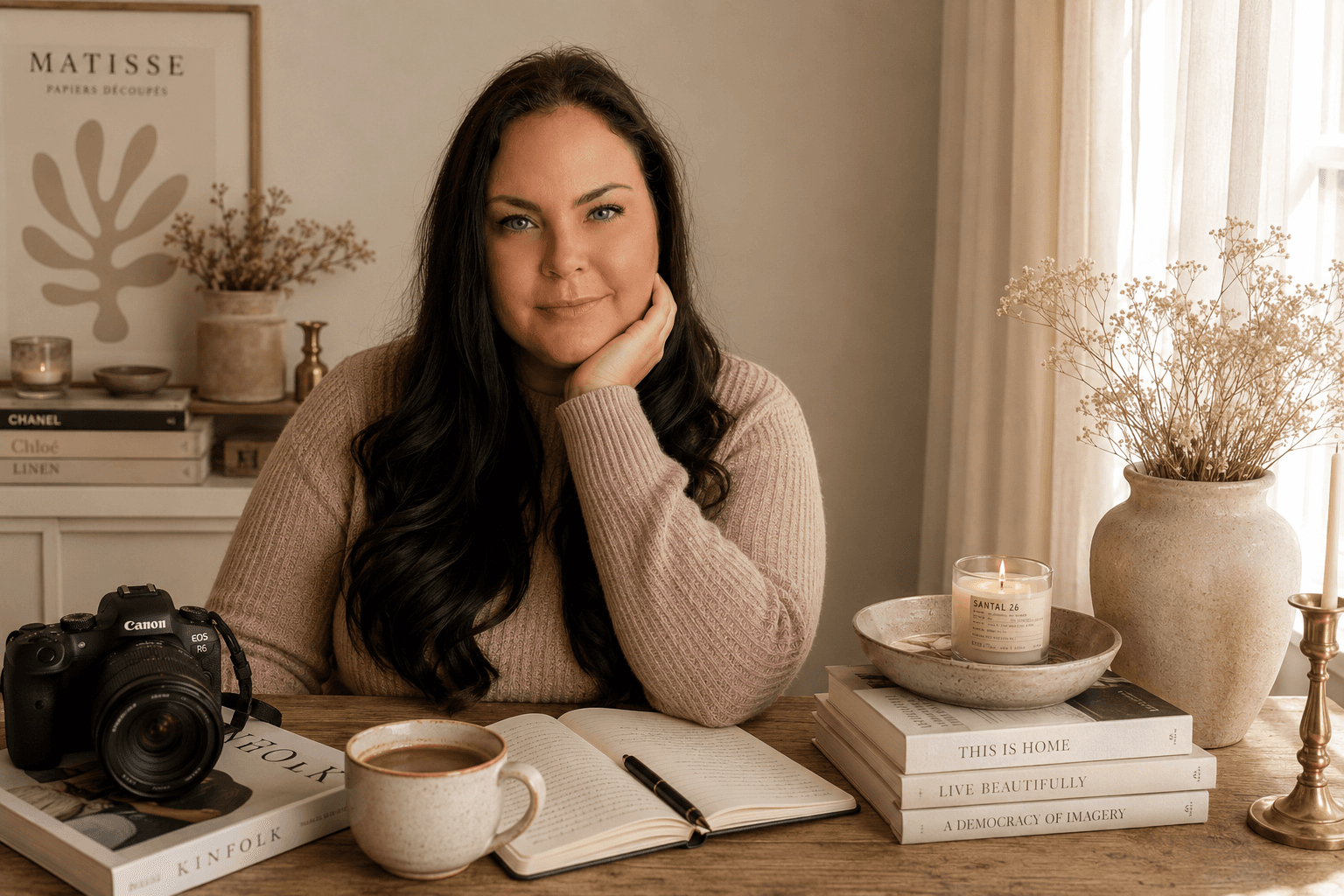 Esther, founder of Essy’s Essentials, resting her chin on her hand at a wooden desk with a camera, an open notebook, coffee, books and a lit candle.