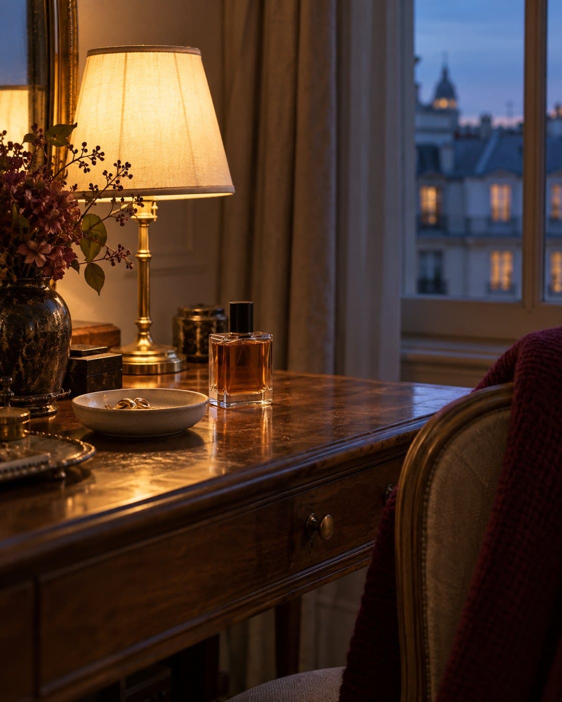 A glass perfume bottle with amber liquid on a polished wooden dressing table beside a lit lamp, a small dish of earrings and dried flowers, with Parisian rooftops at dusk through the window.