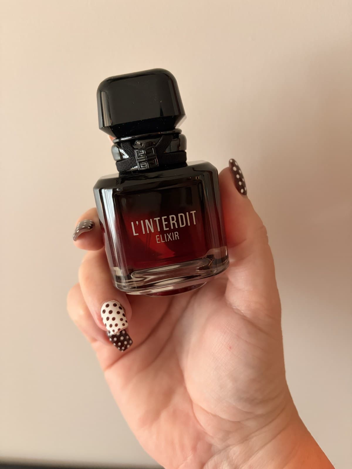 A bottle of Givenchy L'Interdit Elixir eau de parfum held upright in one hand, its dark red liquid visible through the black glass.