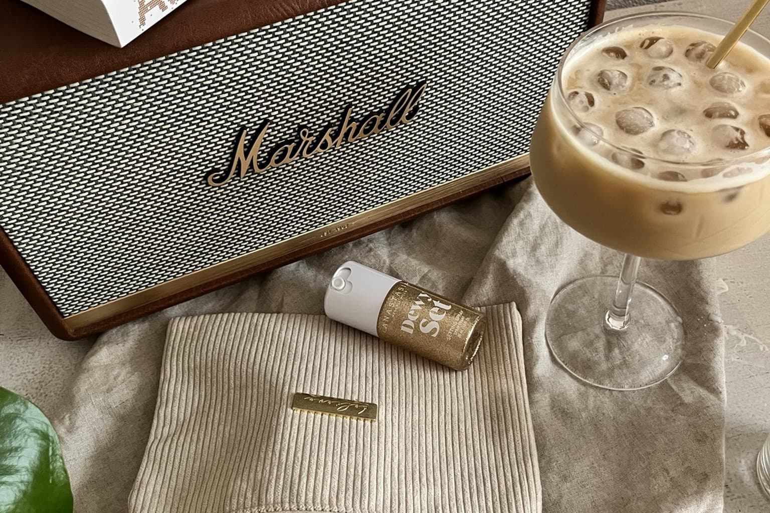 A Marshall speaker, stacked books and an iced coffee beside a corduroy pouch of small skincare and perfume treats