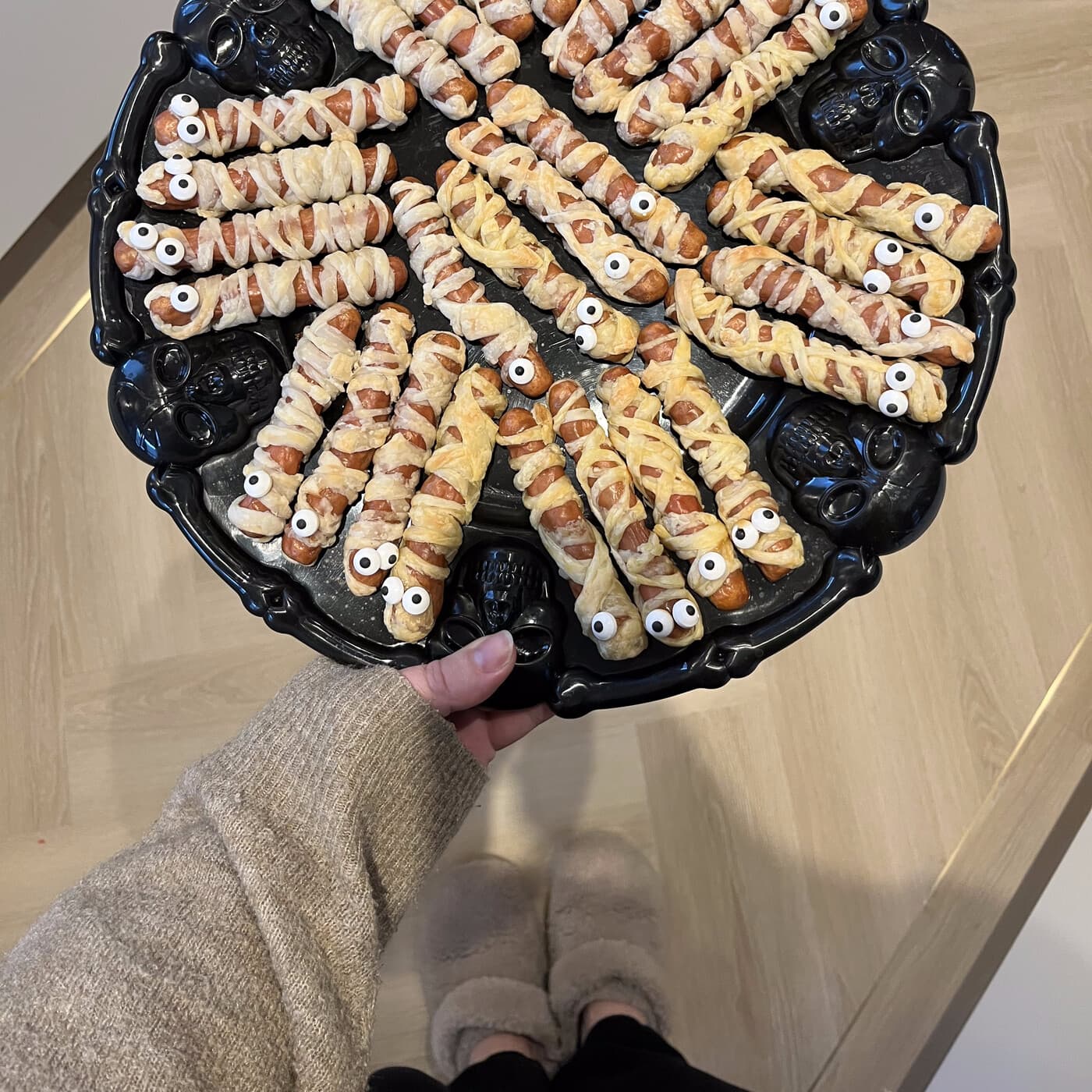 Mummy-wrapped sausage rolls with candy eyes, piled on a skull-shaped platter, held out for guests.