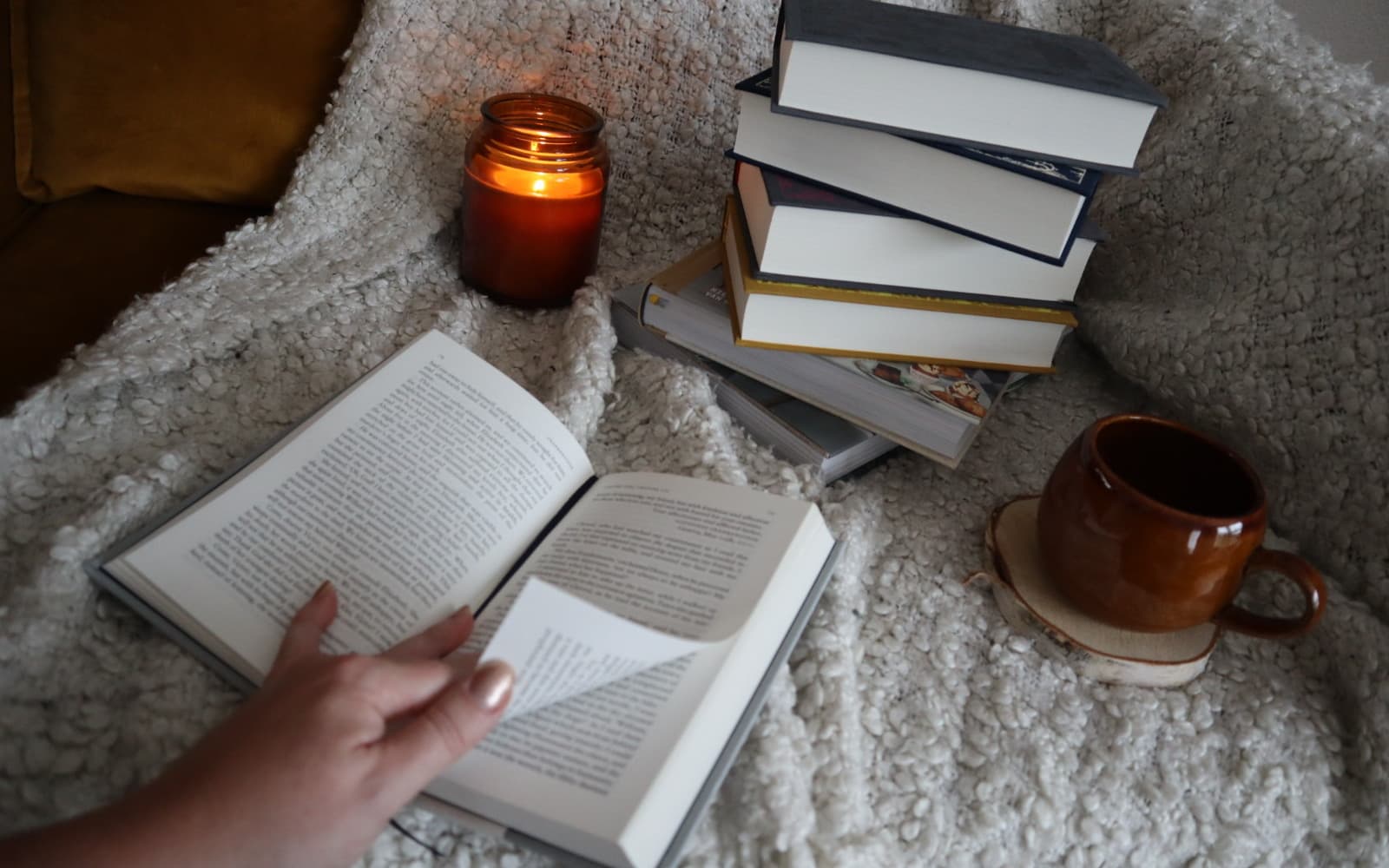 A lit candle and a leaning stack of hardback books beside a hand turning the page of an open book, on a cream knit blanket.