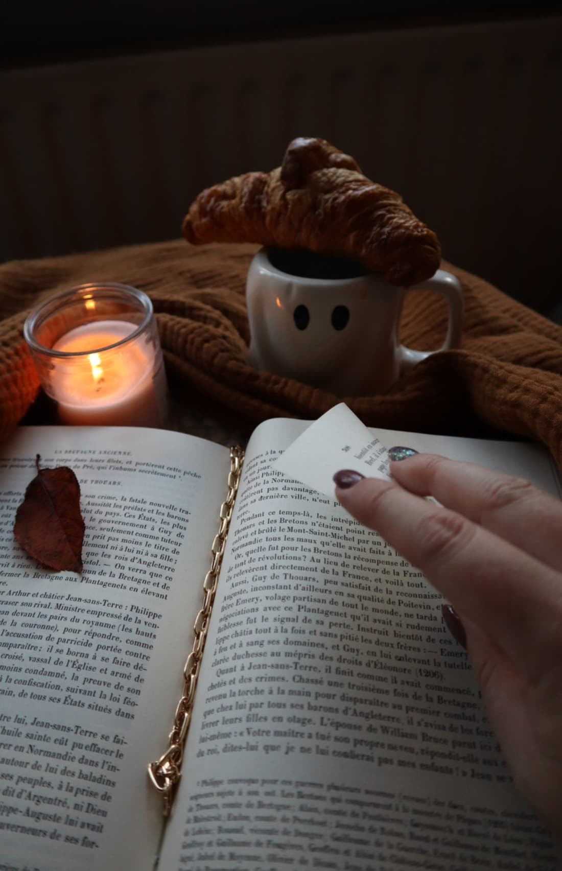 A lit candle and a croissant balanced on a ghost-shaped mug beside an old book with a pressed autumn leaf, in low evening light.