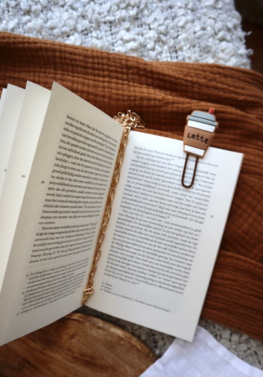 A gold chain bookmark and a small latte-cup bookmark clipped into an open book on a woven blanket.