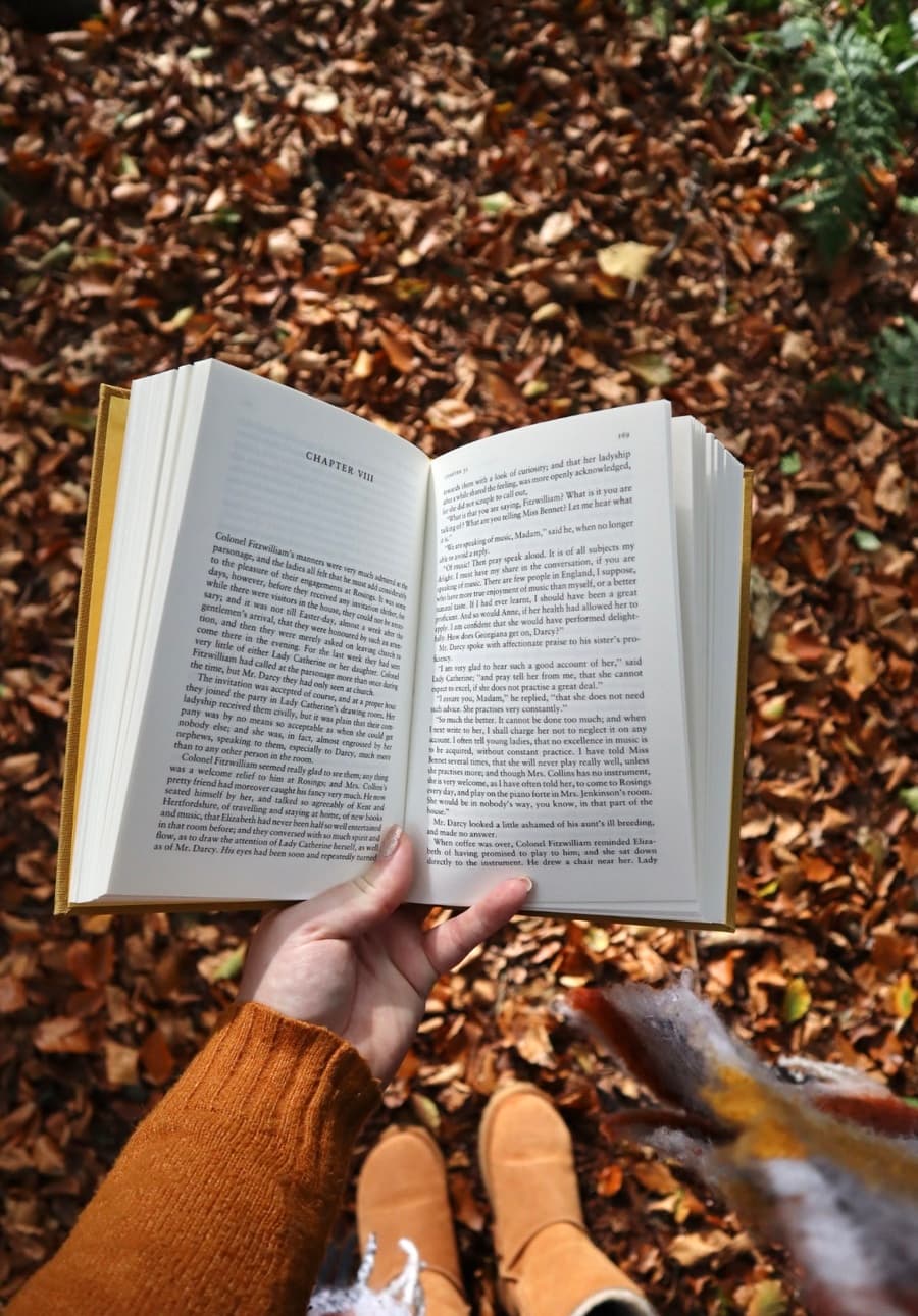 A hand holding an open book outdoors over a bed of autumn leaves, wearing a rust-coloured jumper and suede boots.