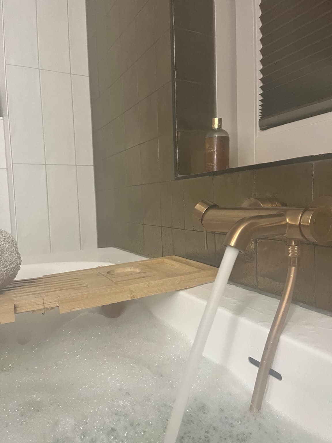 Foaming bath with brushed bronze tap, warm brown tiles and a wooden bath tray.