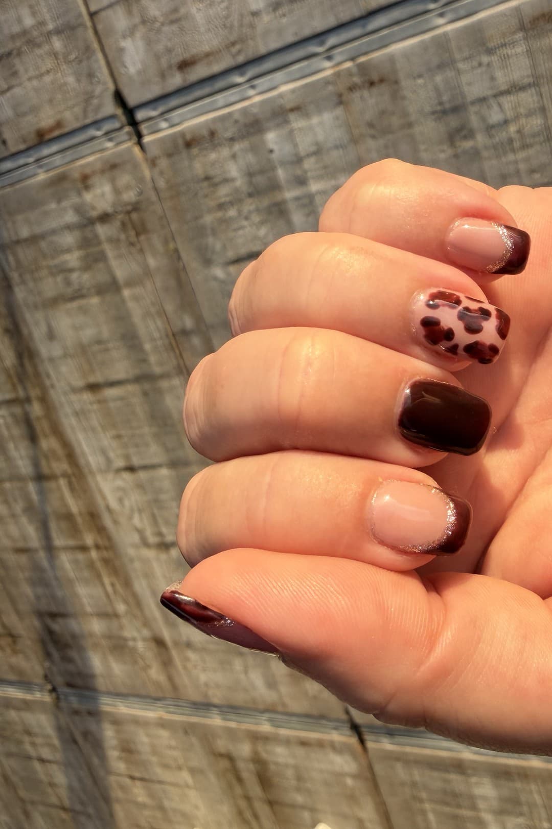 A hand with nails in dark chocolate brown, two with a shimmering gold ombré edge over a nude base, and one accent nail in a brown panther-print pattern.