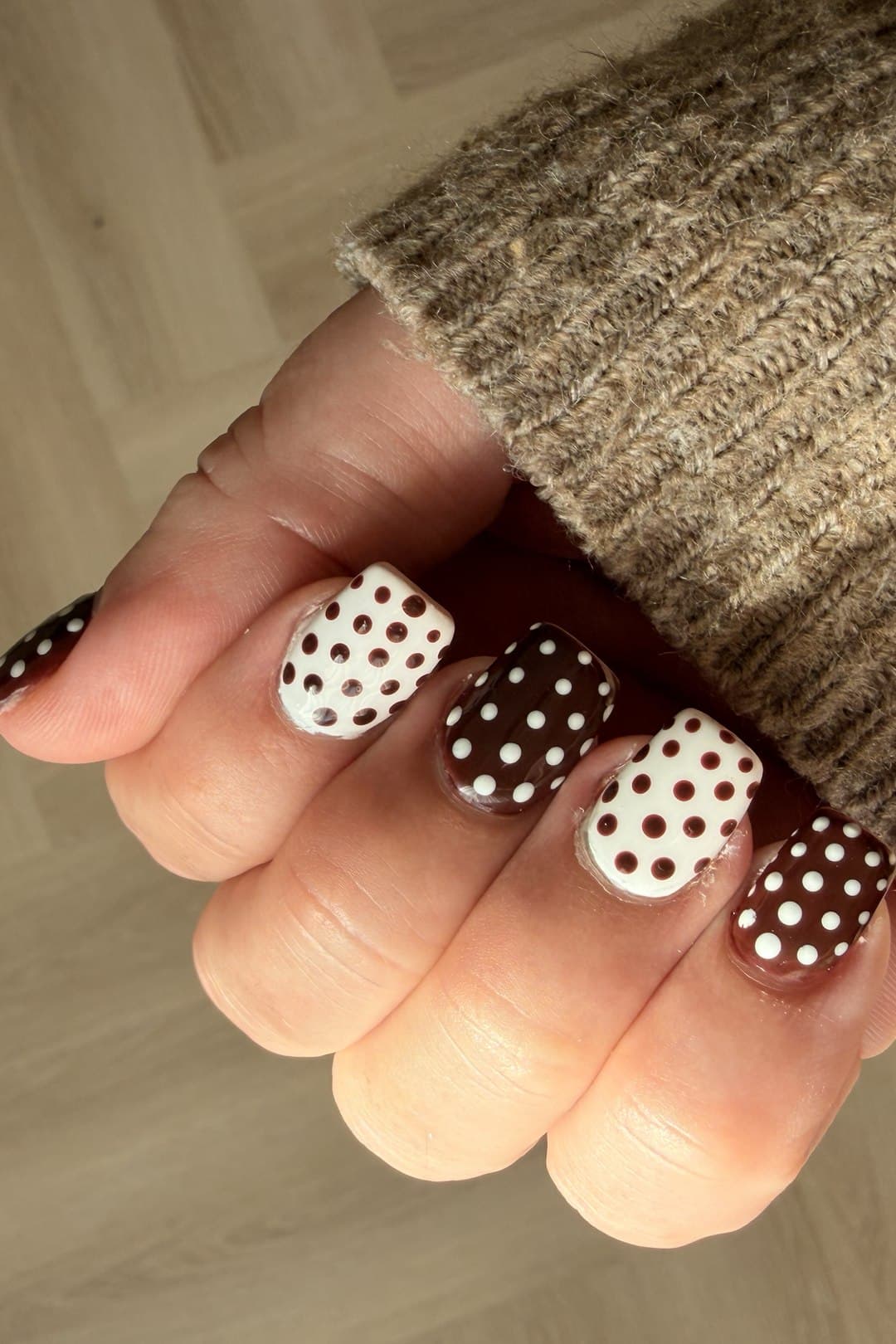 A hand with nails alternating deep cocoa brown and cream, each with a contrasting hand-painted polka dot pattern, resting against a knit sweater sleeve.