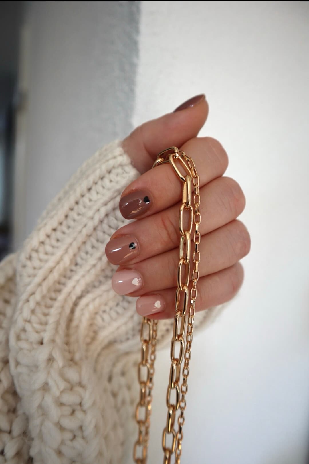 A hand with nails shading from cocoa brown to soft nude, each finished with one tiny heart accent, holding a gold paperclip-chain necklace.