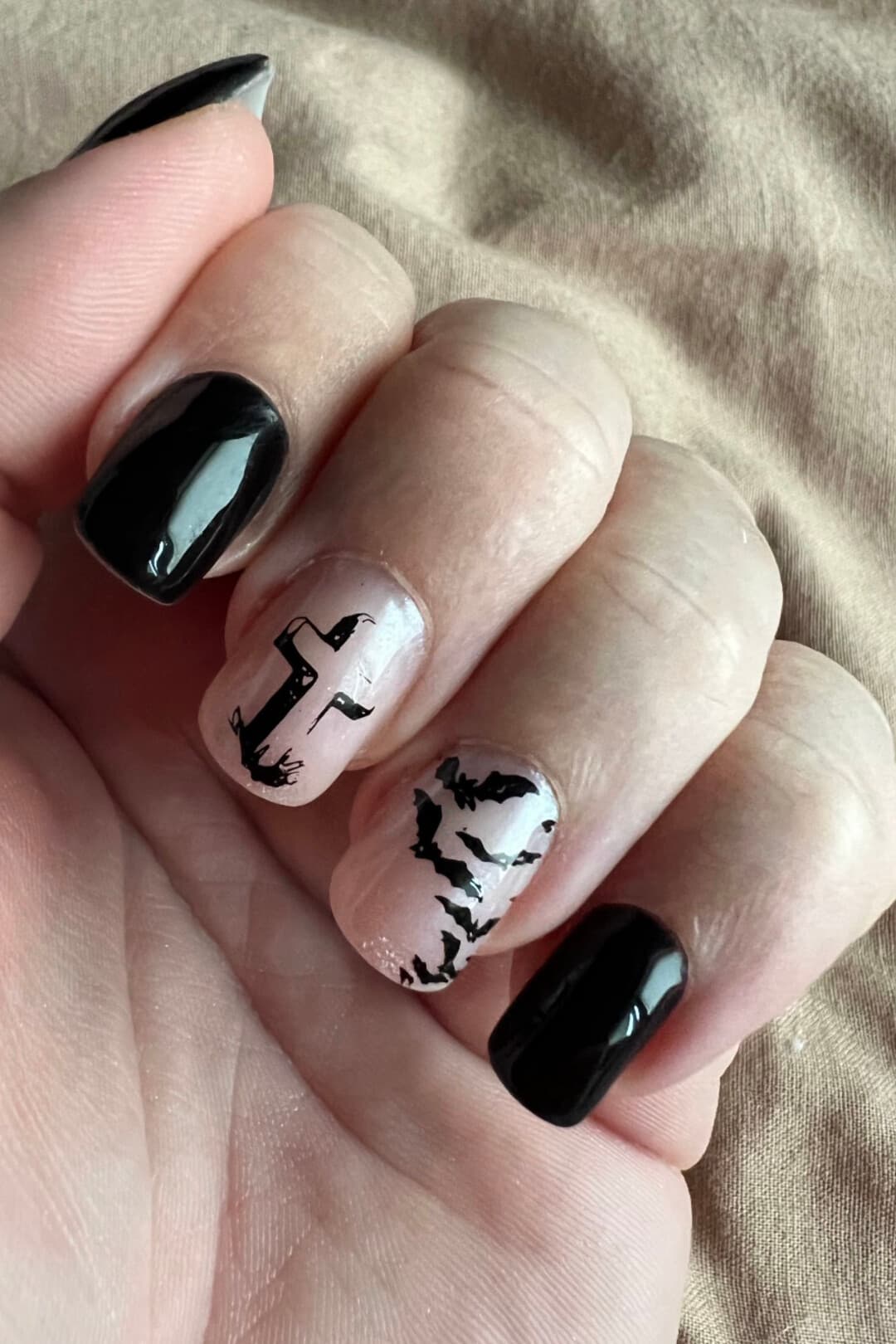A hand with glossy black nails and two nude accent nails, one with a hand-painted cross and one with a flock of hand-painted bats.