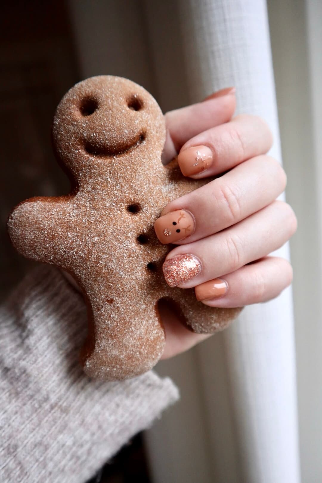 A hand with soft nude nails, one rose gold glitter accent nail and one nail with a small hand-painted reindeer, holding a sugared gingerbread man cookie.