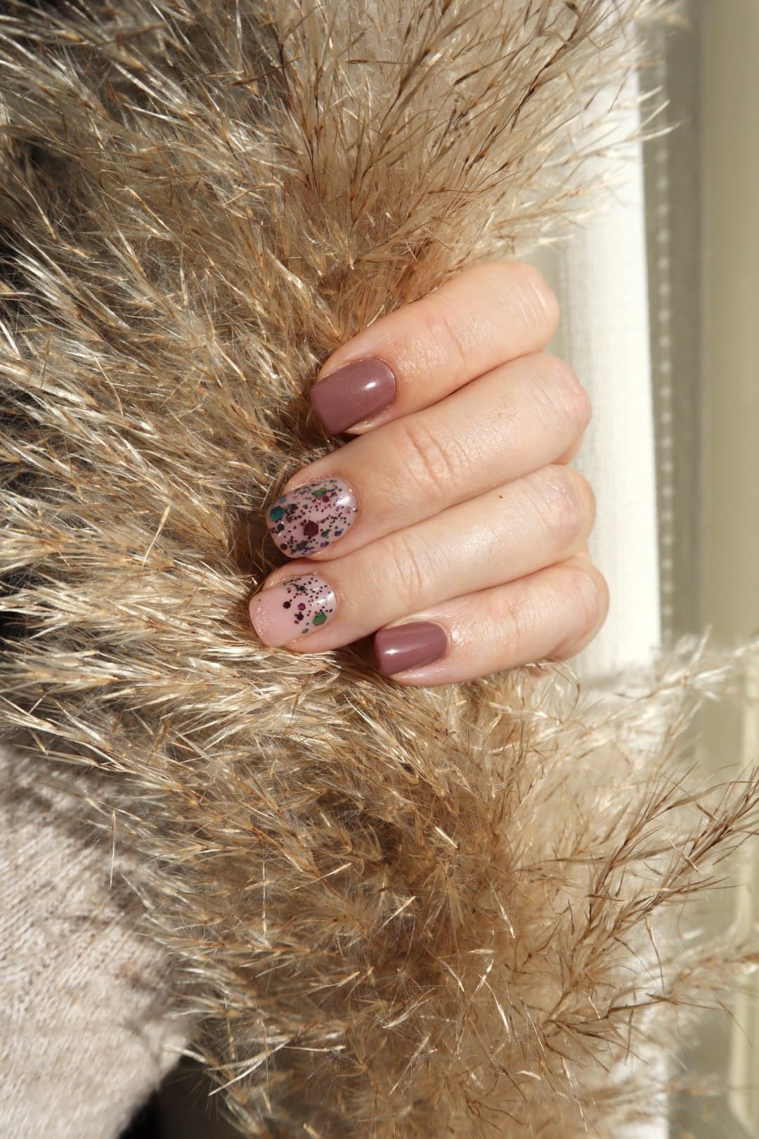 A hand with soft mauve manicured nails and two accent nails carrying scattered dark glitter flecks, resting on a warm faux-fur texture.