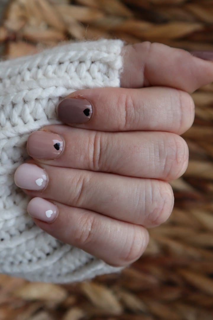 A hand with nails gradating from deeper mauve to soft nude, each finished with one tiny heart accent, resting against a cream knit sleeve.