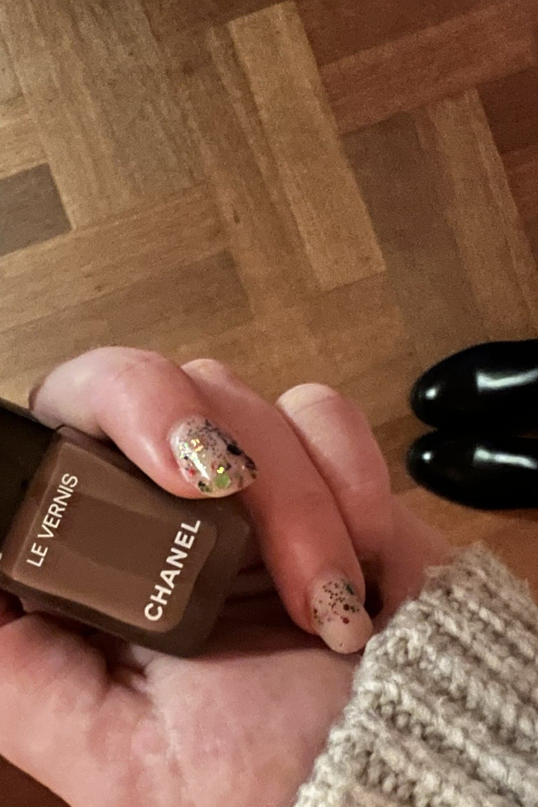 A hand with soft nude nails scattered with colorful confetti glitter, holding a bottle of nail polish, shot on a wood floor.