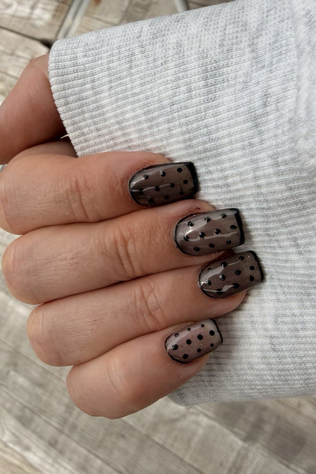 A hand with sheer smoky-brown nails outlined in glossy black, each scattered with black polka dots, resting against a grey knit sleeve.