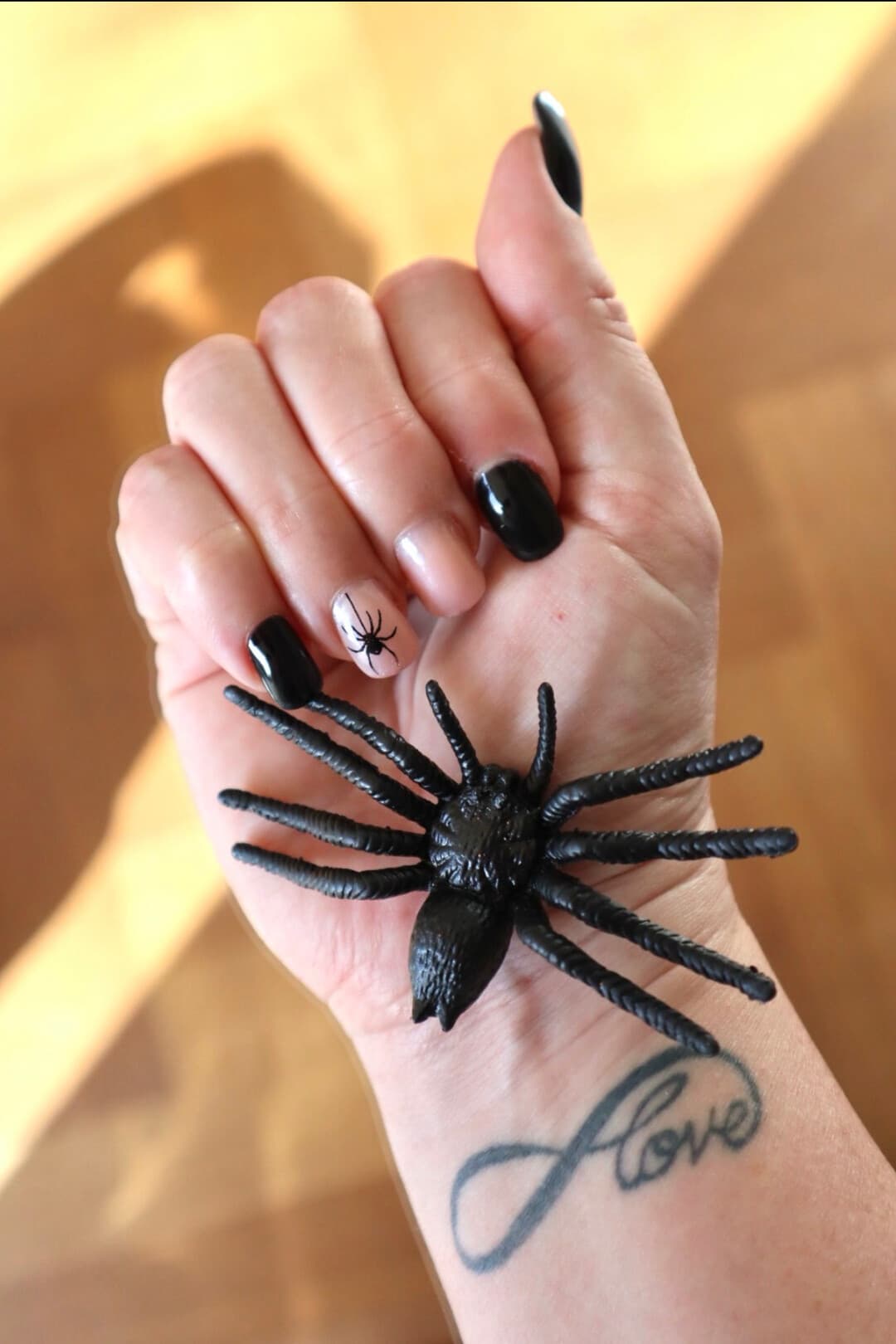 A hand with glossy black nails and one nude accent nail featuring a hand-painted spider, wearing a large black spider ring.