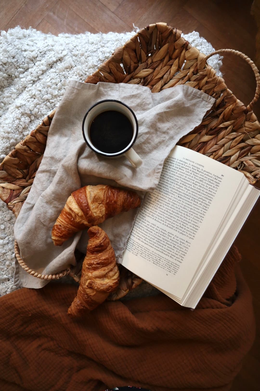 A woven basket holding a French book, two croissants and a black coffee in an enamel mug on a linen cloth, beside a rust-coloured throw.