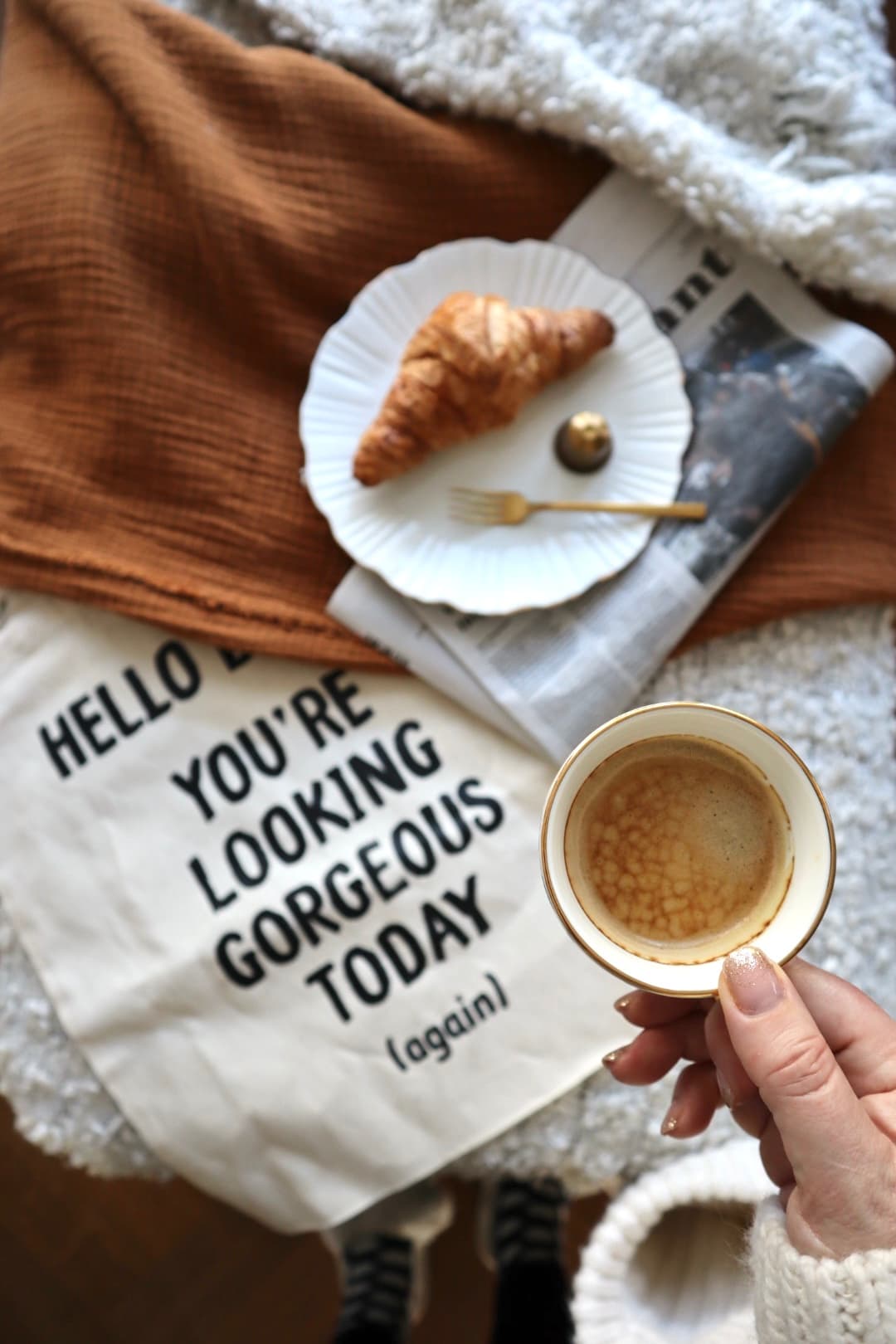 A hand holding a small cup of coffee beside a plate with a croissant and a folded newspaper on a rust-coloured cloth.