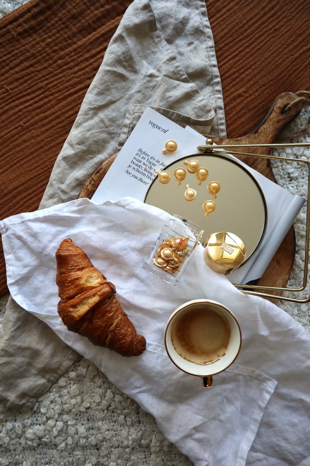 Elizabeth Arden skincare capsules beside coffee, a croissant and a gold mirror in a warm editorial flatlay, with a Vogue.nl print in the background.