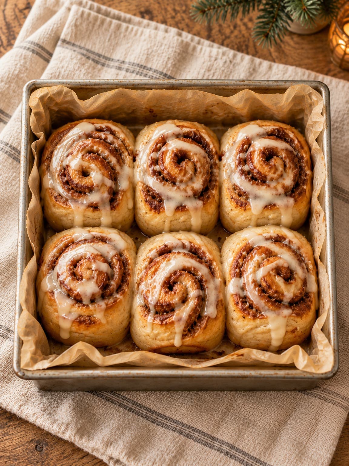Six cinnamon rolls with vanilla icing in a parchment-lined square baking tin.