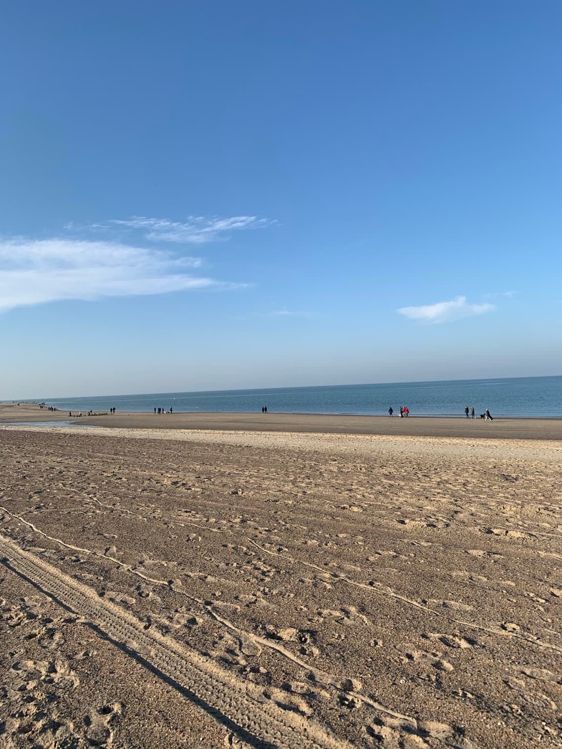 A wide, open stretch of beach under a blue sky, with a few people walking near the distant sea.