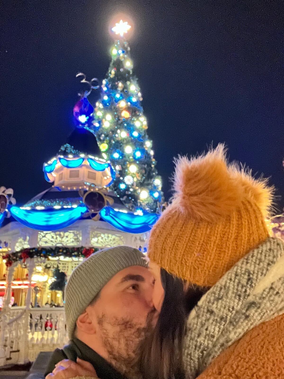 Couple embracing at night beside an illuminated blue Christmas tree and carousel-style building