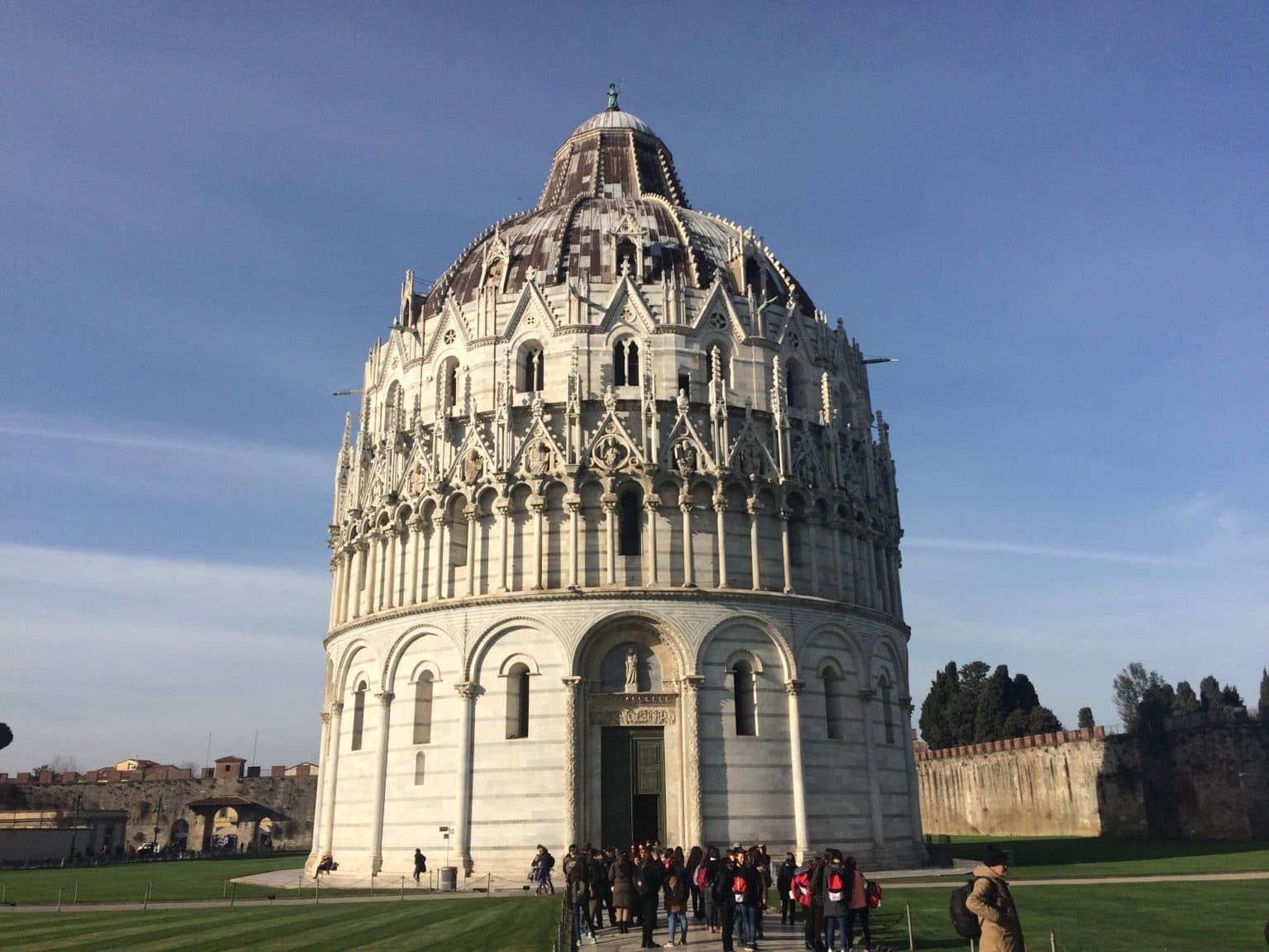 The ornate white-stone Baptistery of Pisa against a deep blue sky, with visitors small at its base