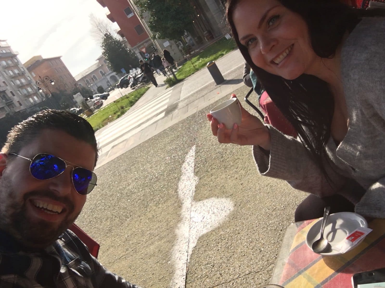 A smiling couple at a sunny pavement café table in Pisa, one holding a small white espresso cup