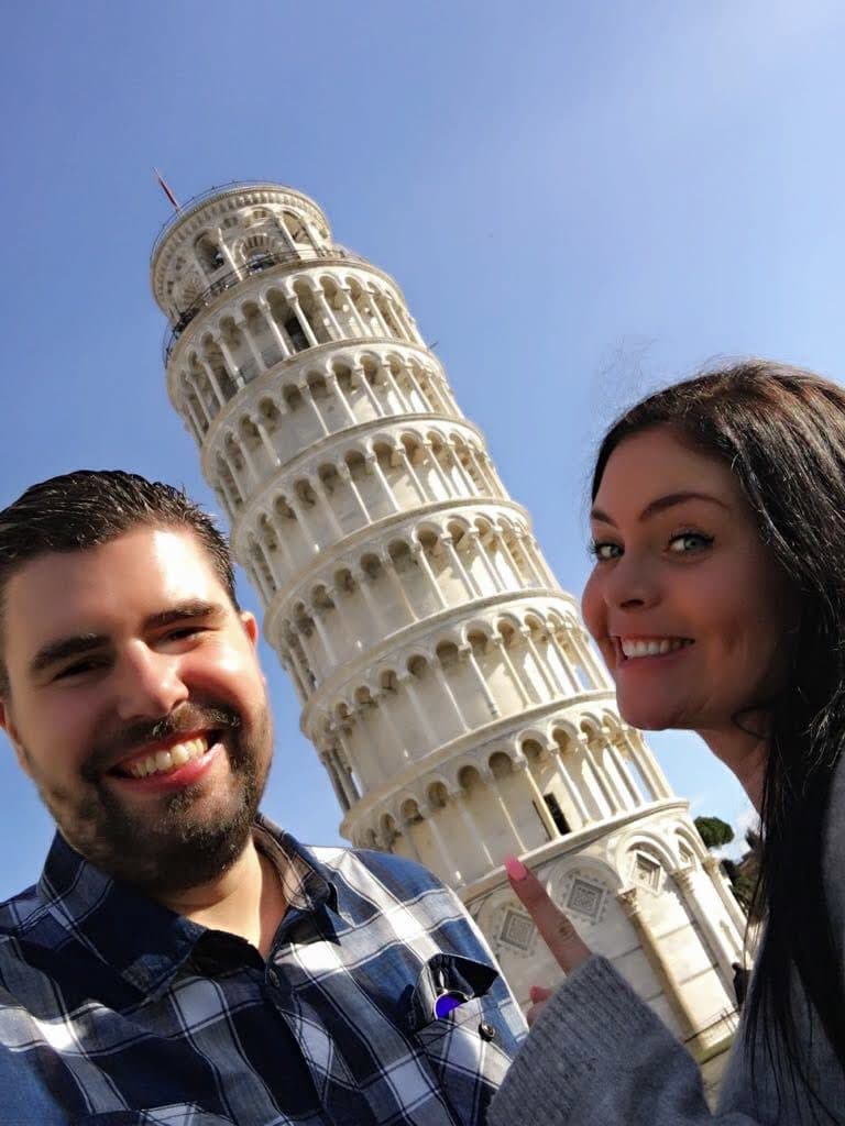A couple smiling in a selfie in front of the Leaning Tower of Pisa against a clear blue sky