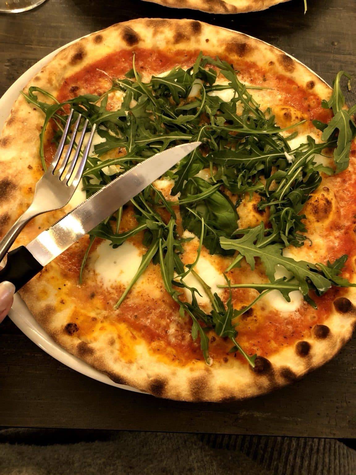 A blistered-crust pizza topped with tomato, mozzarella and a heap of rocket, with a knife and fork on the plate