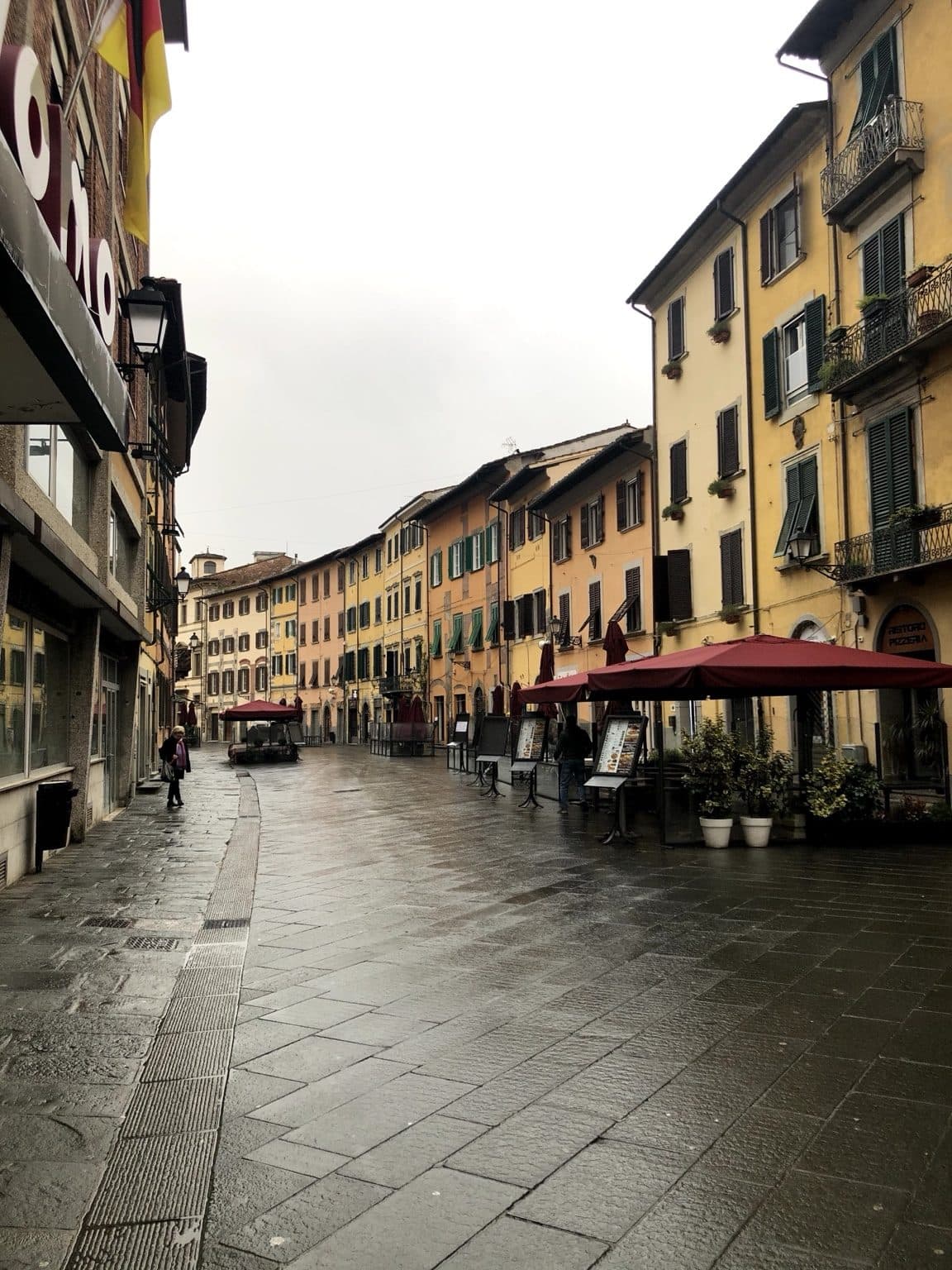 A wet Pisa street lined with ochre and yellow facades, dark shiny paving and folded restaurant umbrellas under a grey sky