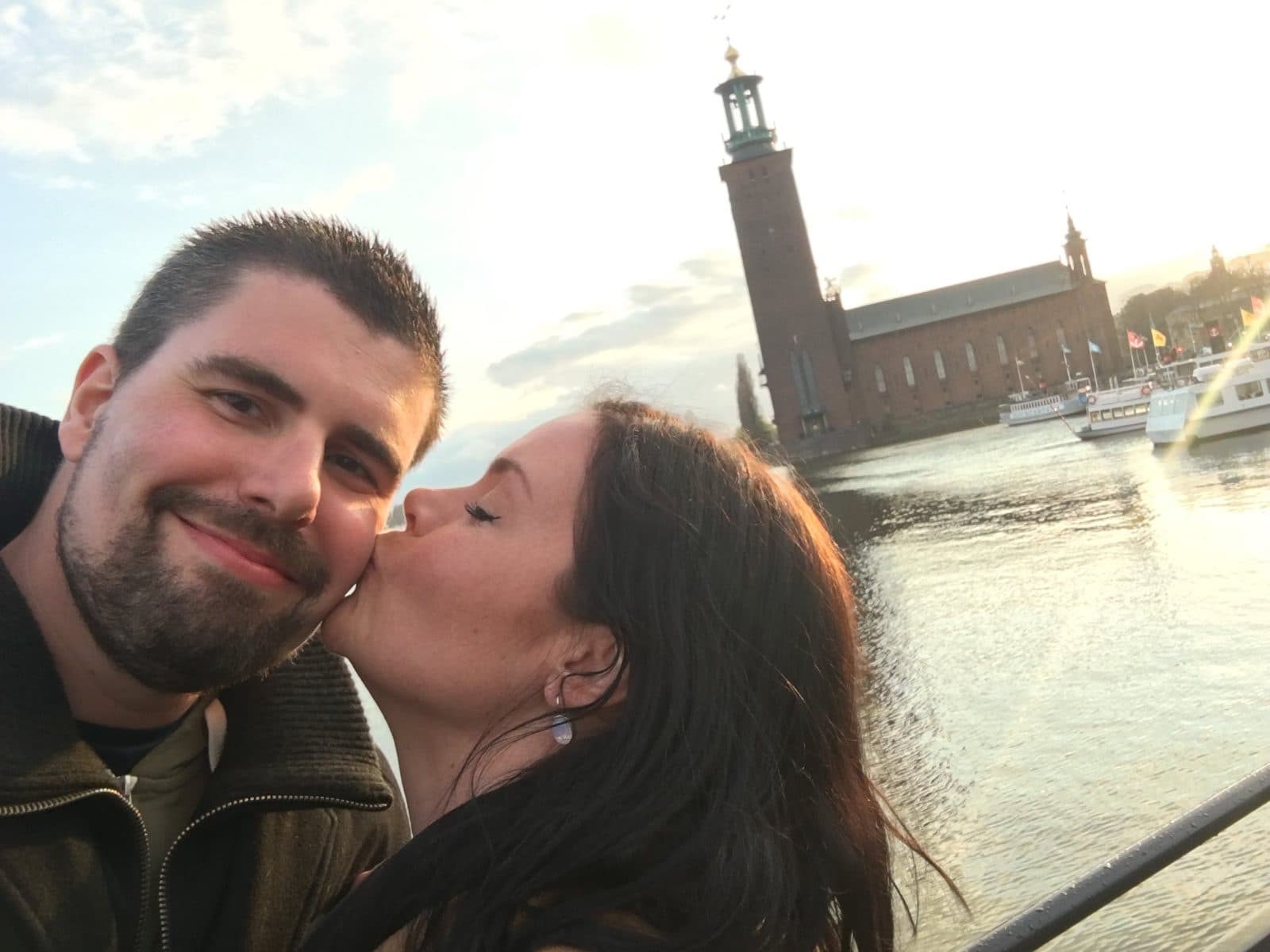 A couple in a close selfie, one kissing the other on the cheek, with Stockholm City Hall’s brick tower and the water behind them in low sunlight