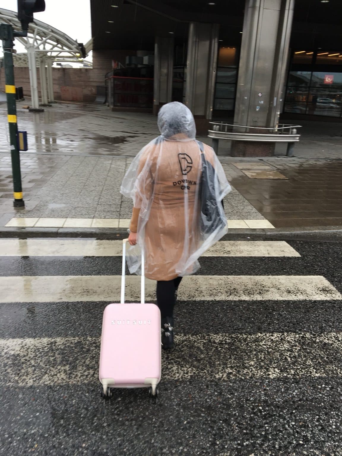 A person in a clear rain poncho pulling a pink suitcase across a wet zebra crossing in Stockholm, seen from behind