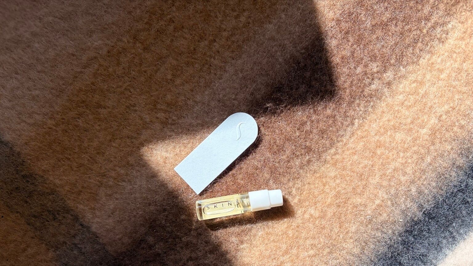 Skins perfume sample and embossed scent strip on brown fabric in sunlight.