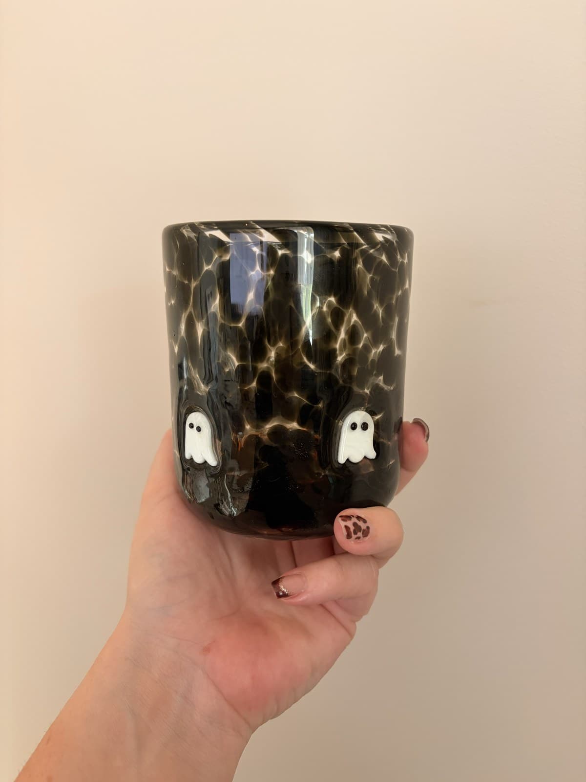 A dark tortoiseshell-patterned glass tumbler with two small white ghosts on its side, held in one hand.