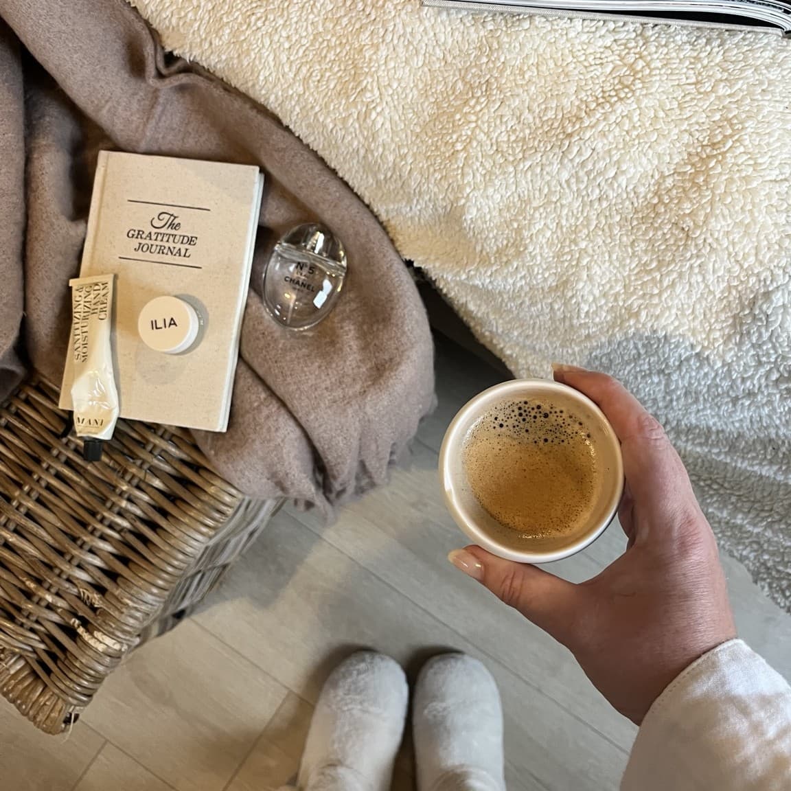 A hand holding a cup of coffee beside a gratitude journal, hand cream and perfume on a cosy blanket, with slippers on the floor below