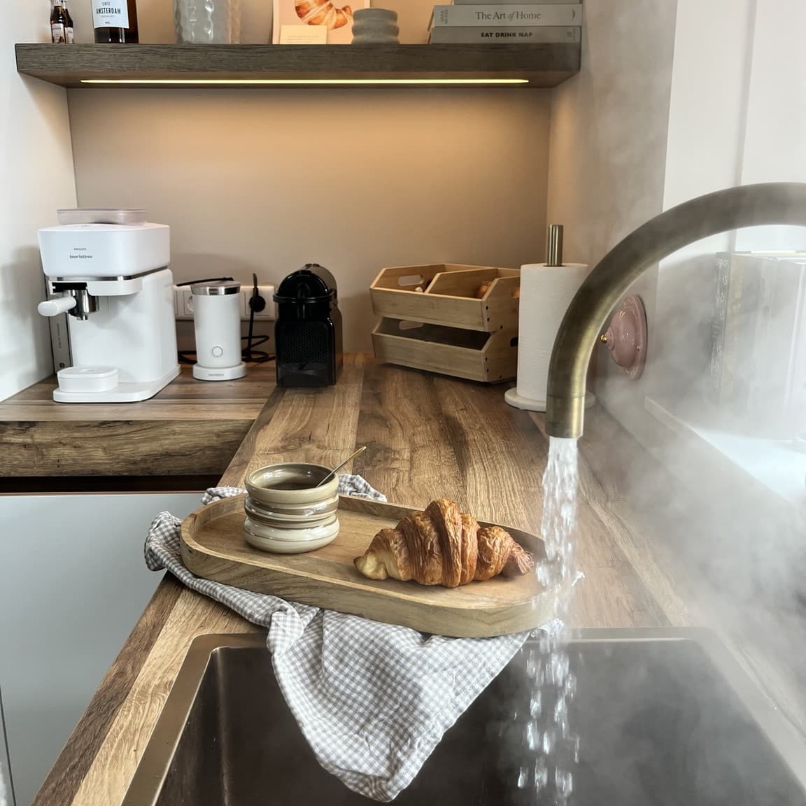 A wooden tray with coffee and a croissant on a kitchen counter, steam rising from a running tap beside it