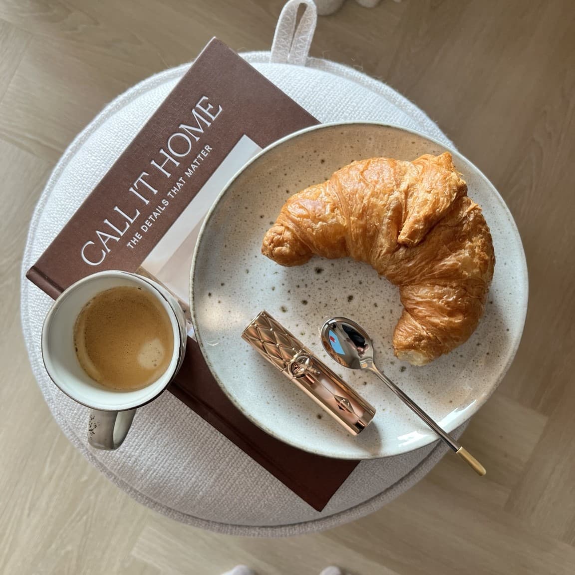 A book called "Call It Home", a cup of coffee, a croissant and a rose-gold lipstick arranged on a plate, resting on a round ottoman