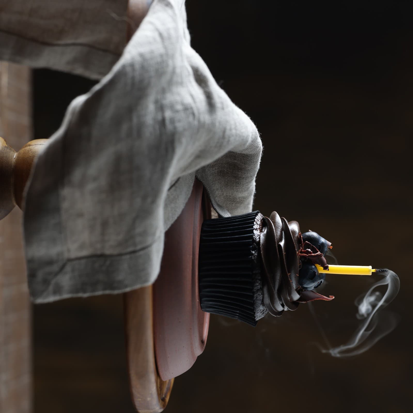 A just-blown-out candle with a lit match resting on it, a thin curl of smoke rising, a linen cloth draped behind it