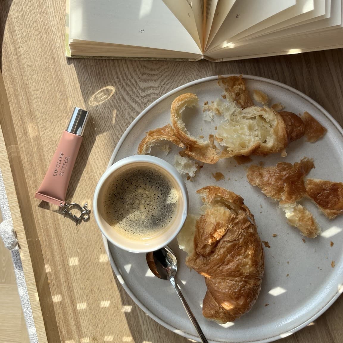 A cracked-open croissant and a cup of coffee on a plate beside an open book and a lip balm, in warm afternoon light