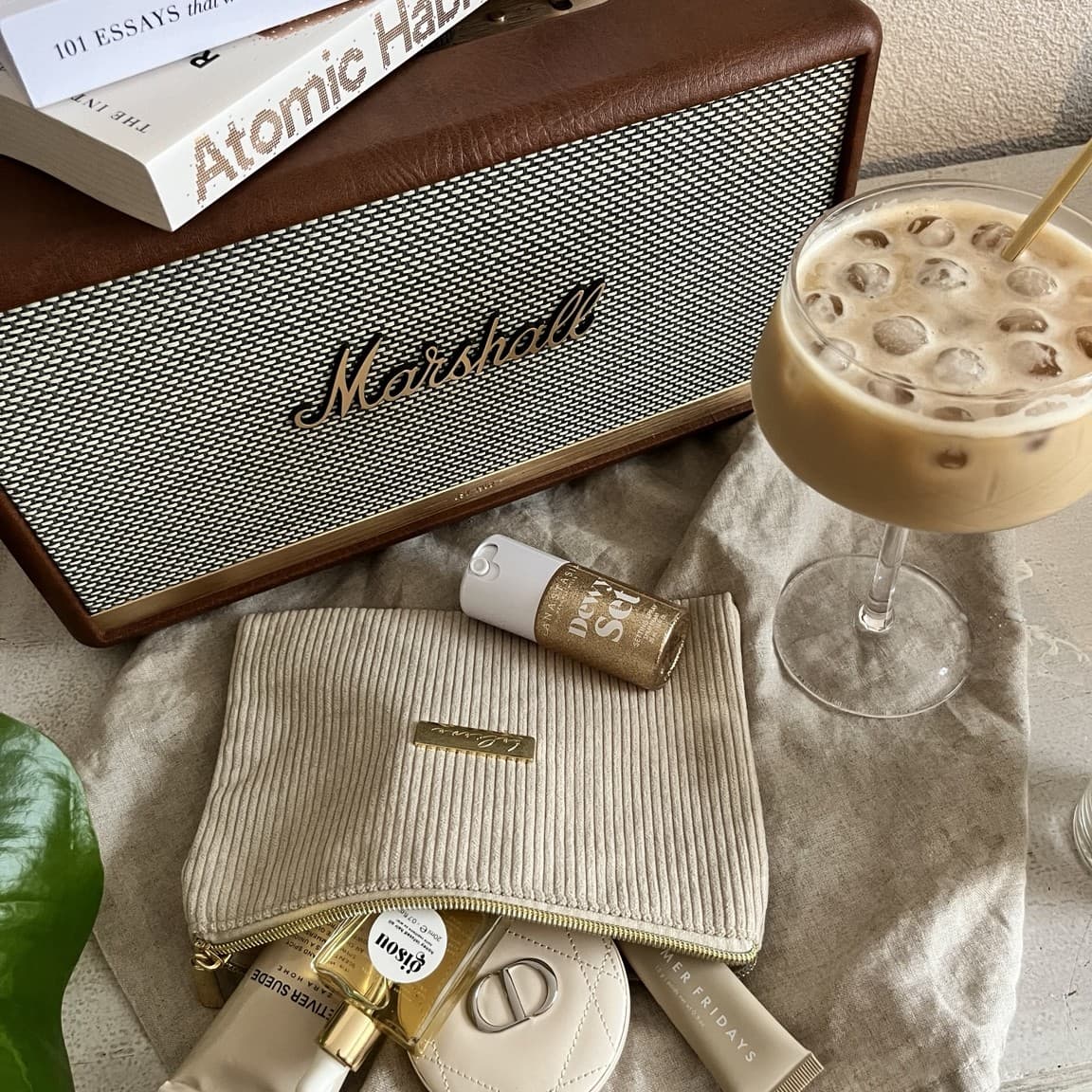 A Marshall speaker and stacked books beside an iced coffee and a small pouch of beauty products on a linen cloth