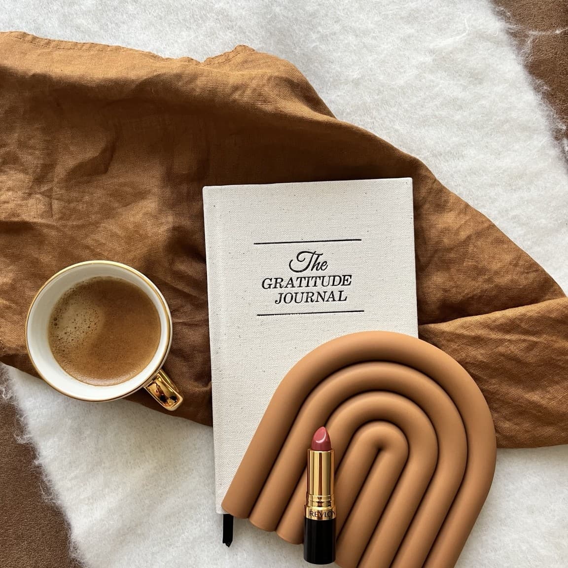 A cup of coffee and a lipstick resting on "The Gratitude Journal" book, on a caramel-coloured linen throw