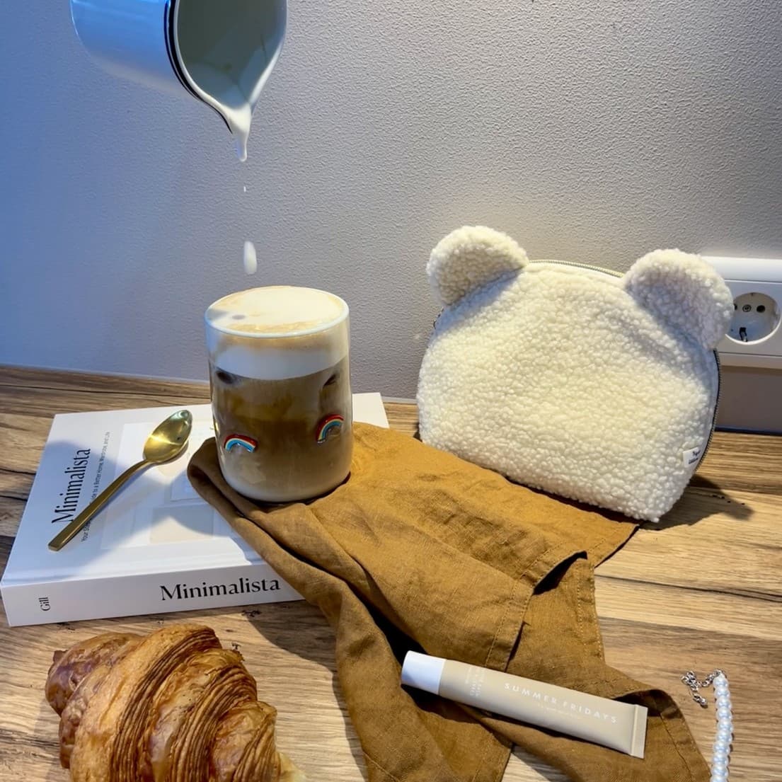 Milk being poured into a rainbow-decorated latte glass beside a croissant, a teddy-bear-shaped pouch and a strand of pearls