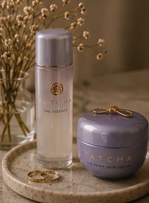 Tatcha The Essence bottle and The Dewy Skin Cream jar beside gold rings and dried flowers.