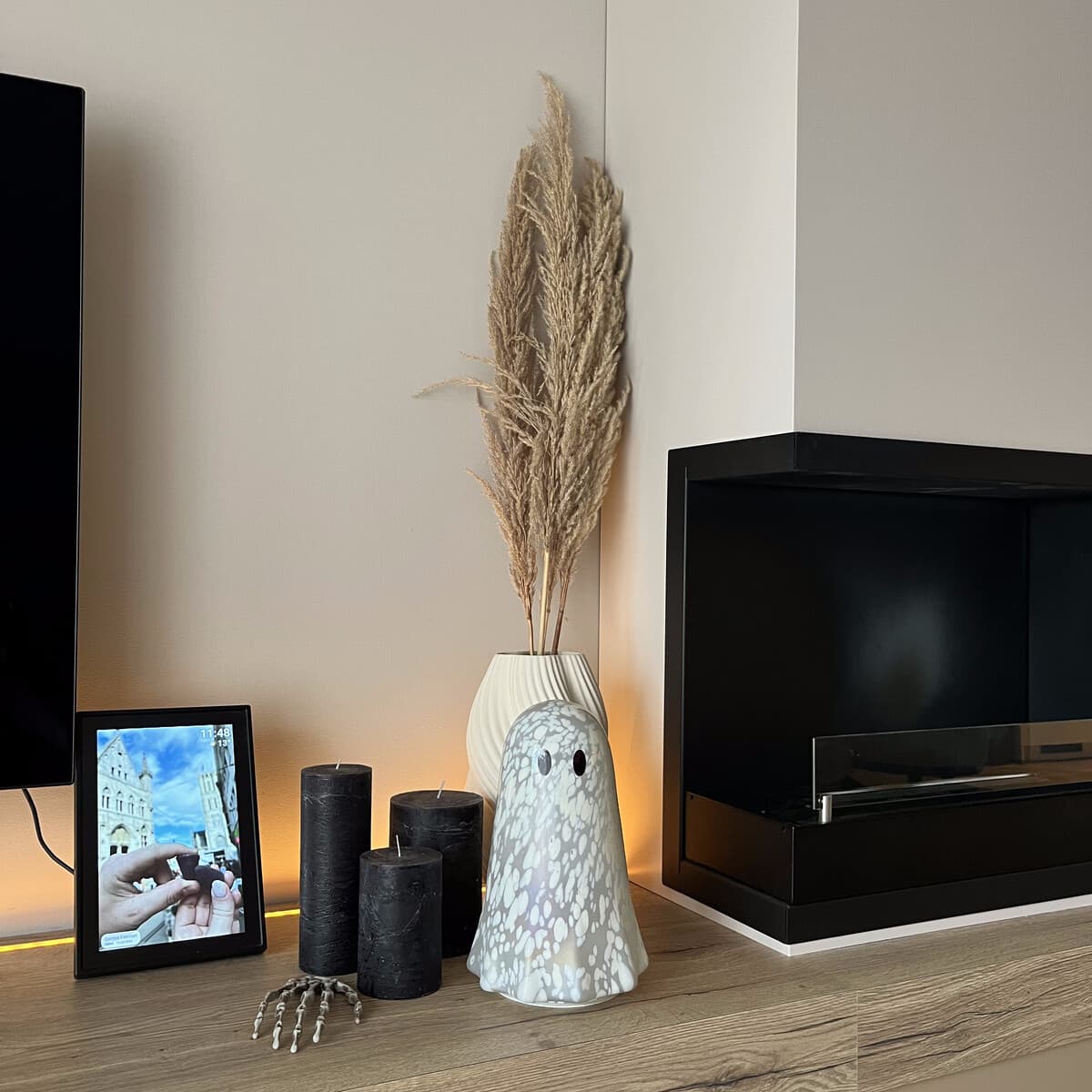 A glowing ghost lamp, three black pillar candles and a small skeletal hand ornament, arranged on a console table beside a vase of pampas grass.