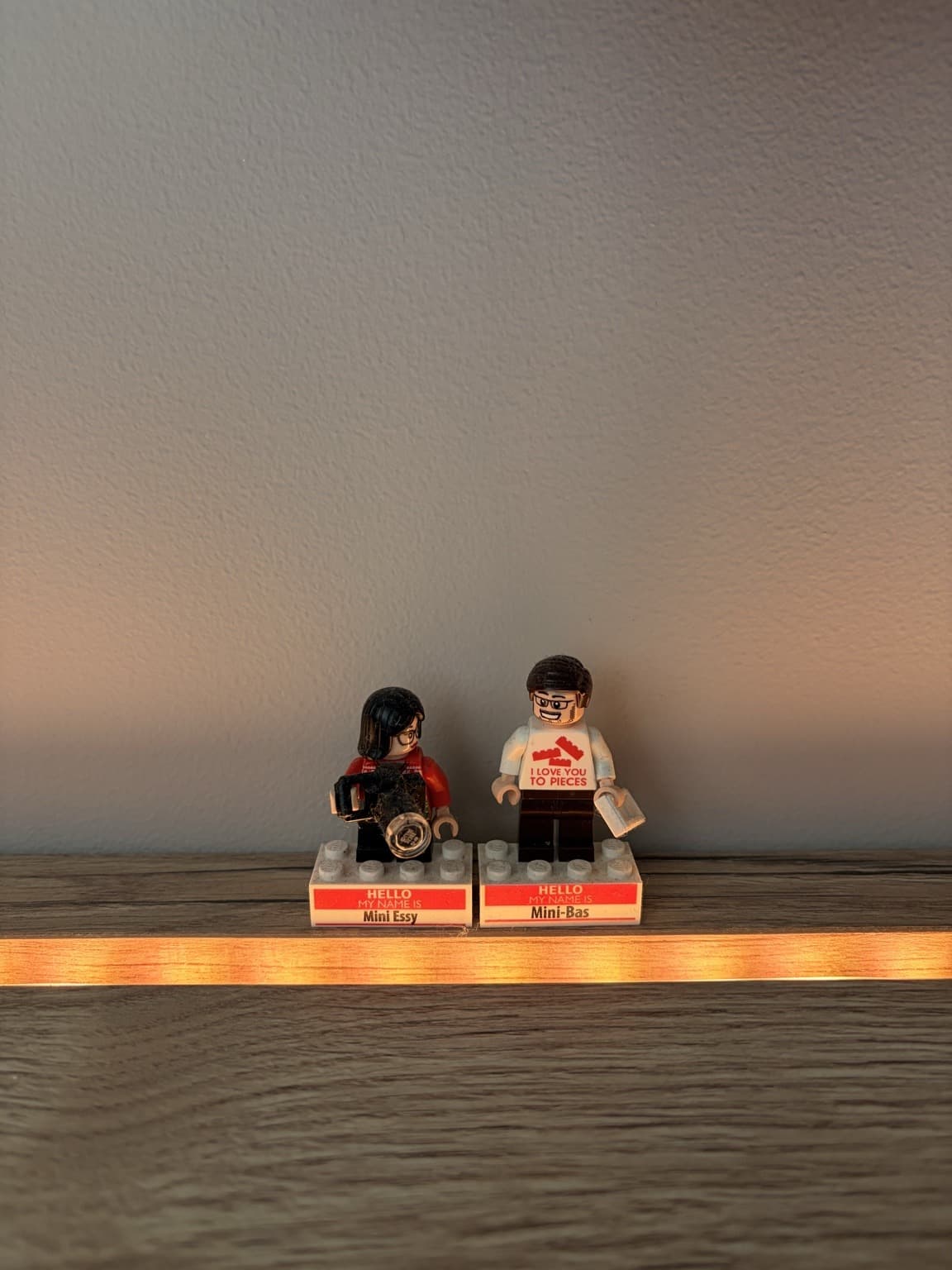 Two Lego minifigures labelled Mini Essy and Mini Bas on a wooden shelf.