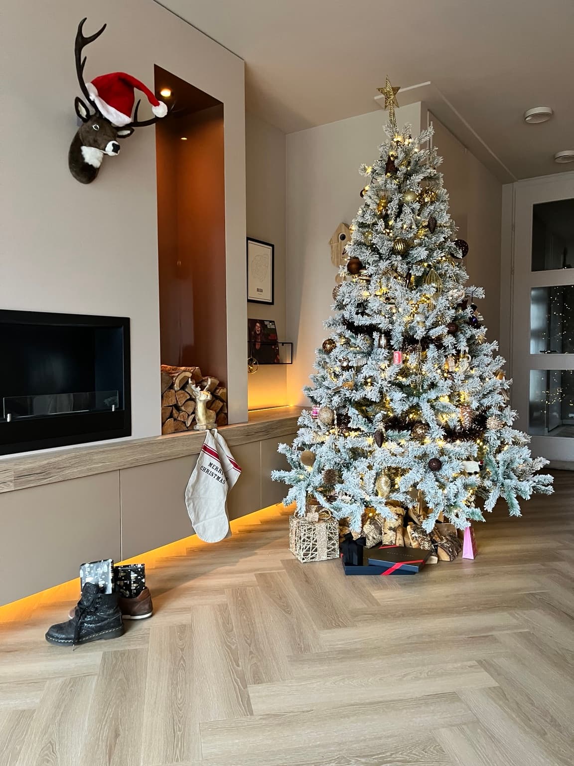 A tall snow-covered Christmas tree with warm lights and gold and brown ornaments beside the fireplace in a high-ceilinged living room, with a few wrapped presents underneath.