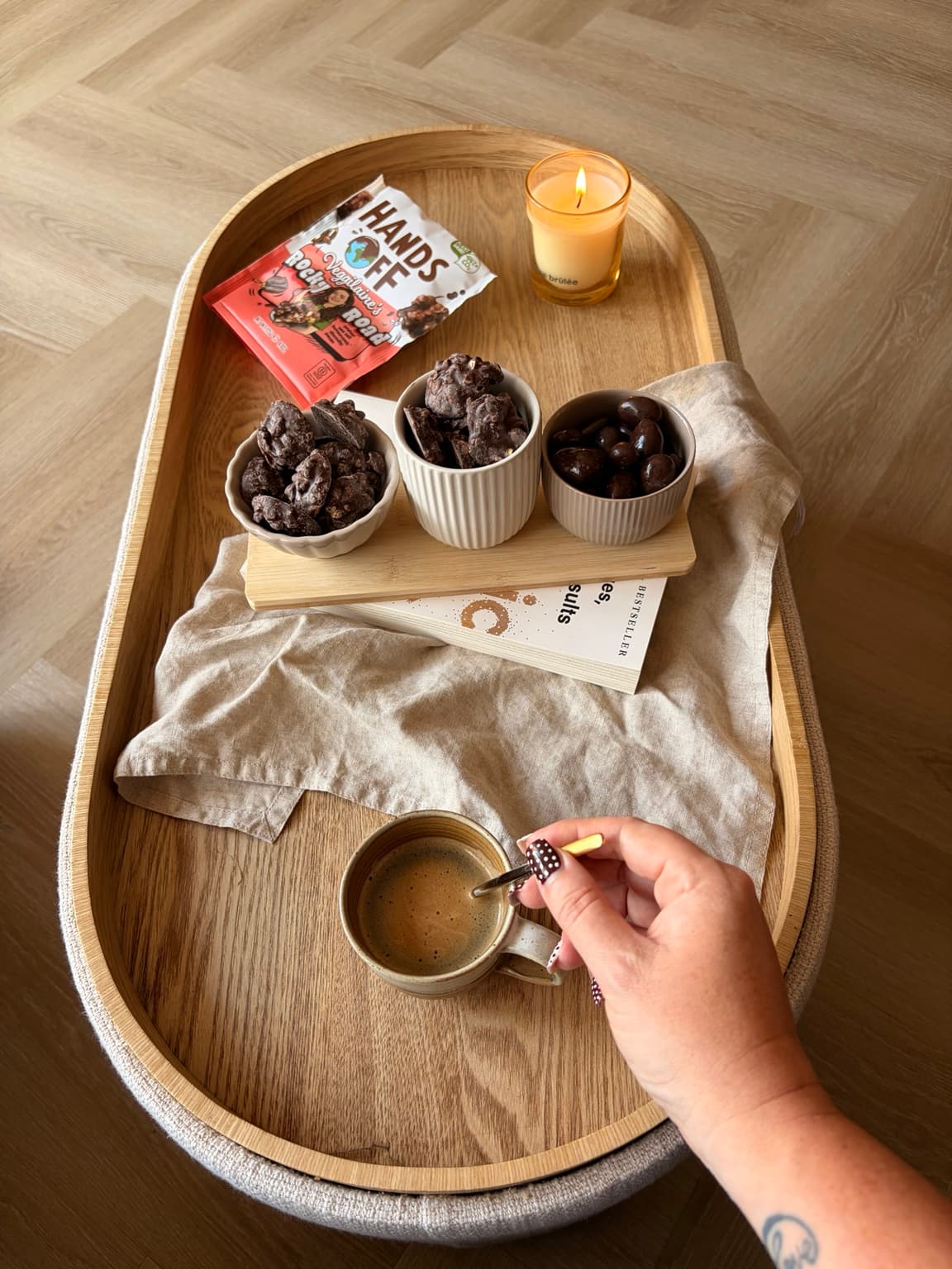 A hand holding a cup of coffee beside a wooden tray with three small bowls of chocolate bites and a Hands Off Rocky Road packet.