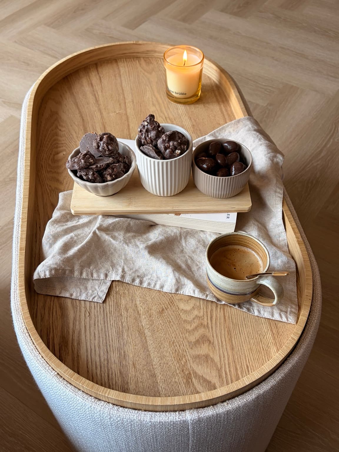 Three small bowls of chocolate bites on a wooden board, with a lit candle and a cup of coffee on a linen napkin.