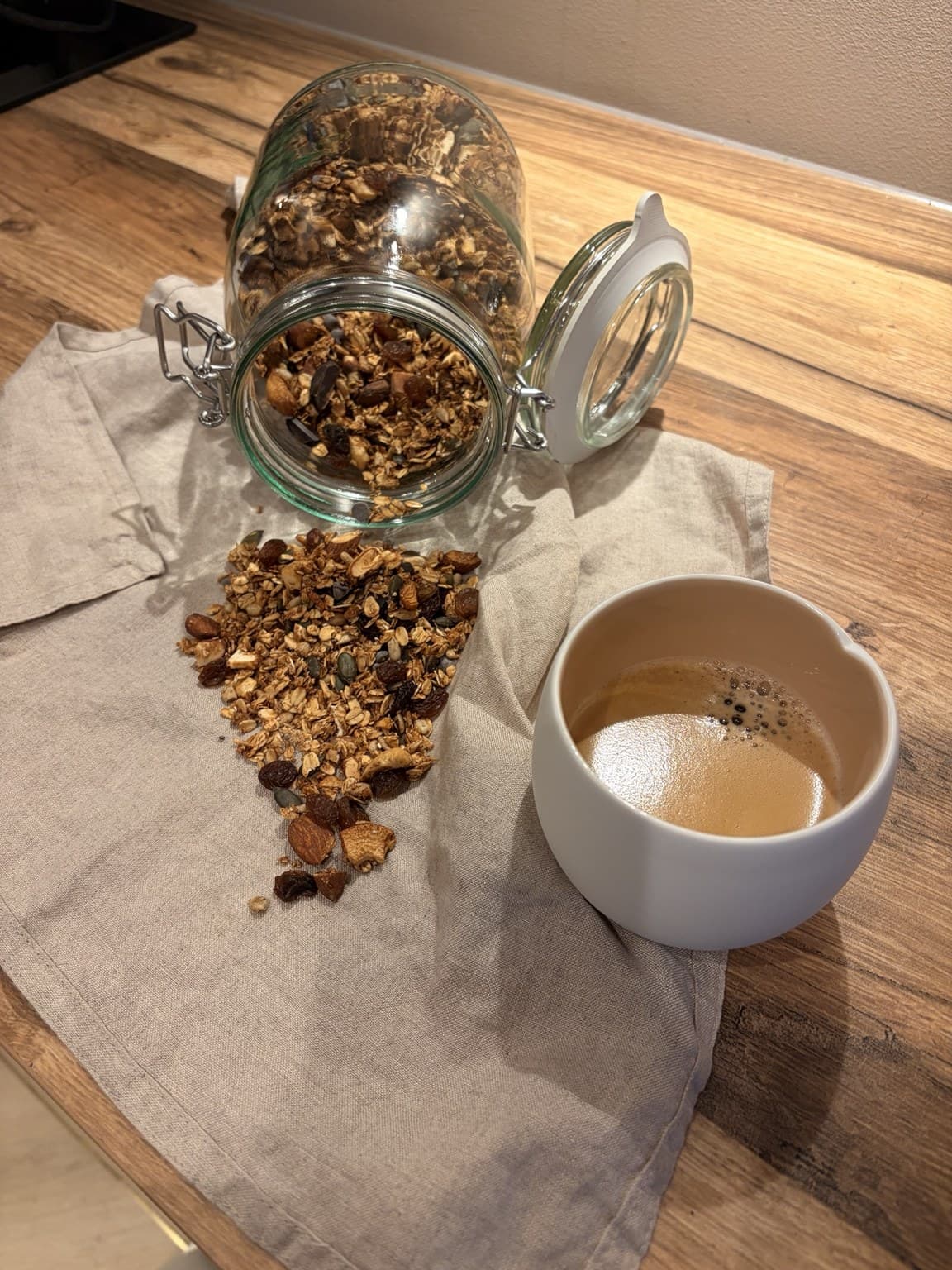 An open jar of granola spilled onto a linen cloth beside a cup of coffee on a wooden table